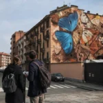 Due ospiti osservano il murale di farfalle The Ode of Collapse di Mantra nel quartiere Aurora a Torino durante un itinerario a piedi.